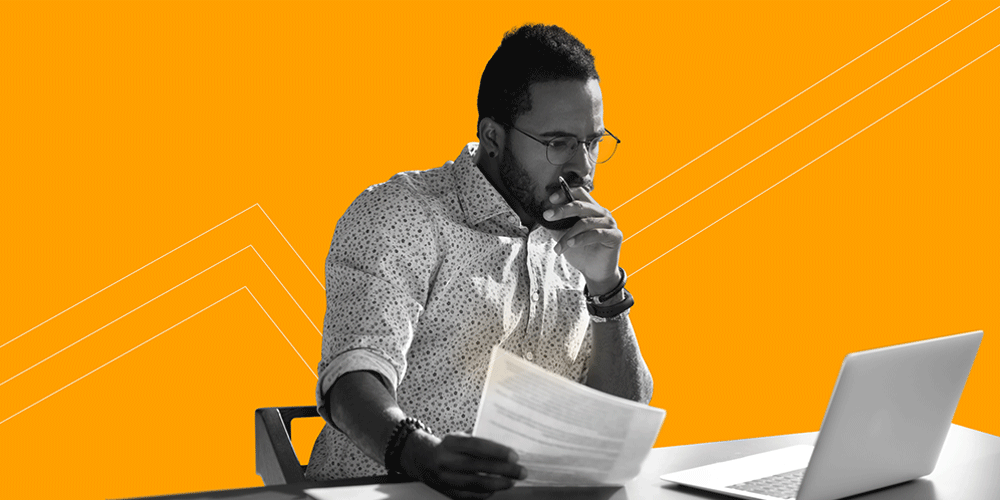 Man looking intently at a laptop while holding a pencil to his mouth with one hand and some papers in the other